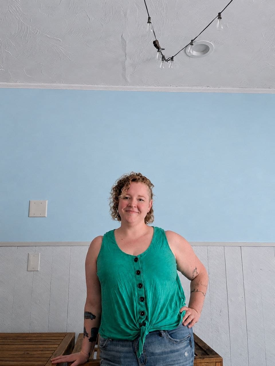 Smiling woman with curly hair wearing a green tank top against a blue wall.