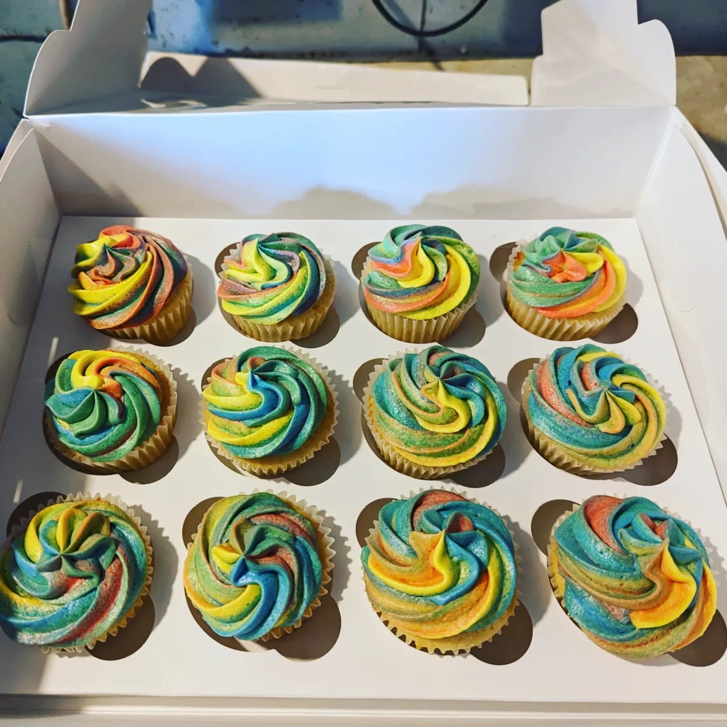 A dozen cupcakes with vibrant, multi-colored rainbow frosting swirls inside a white bakery box.