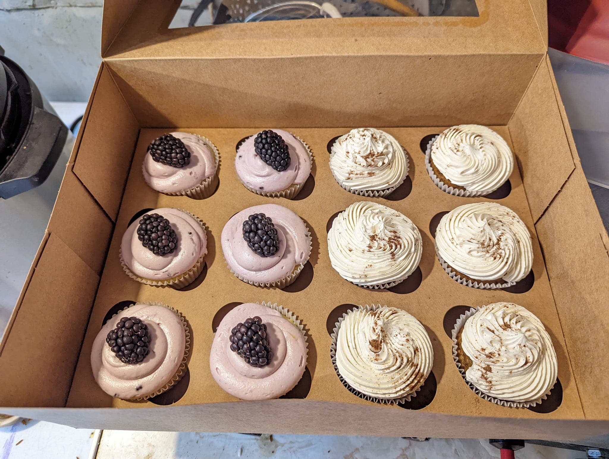 Twelve cupcakes in a box, half with blackberry frosting and half with white cinnamon swirls.