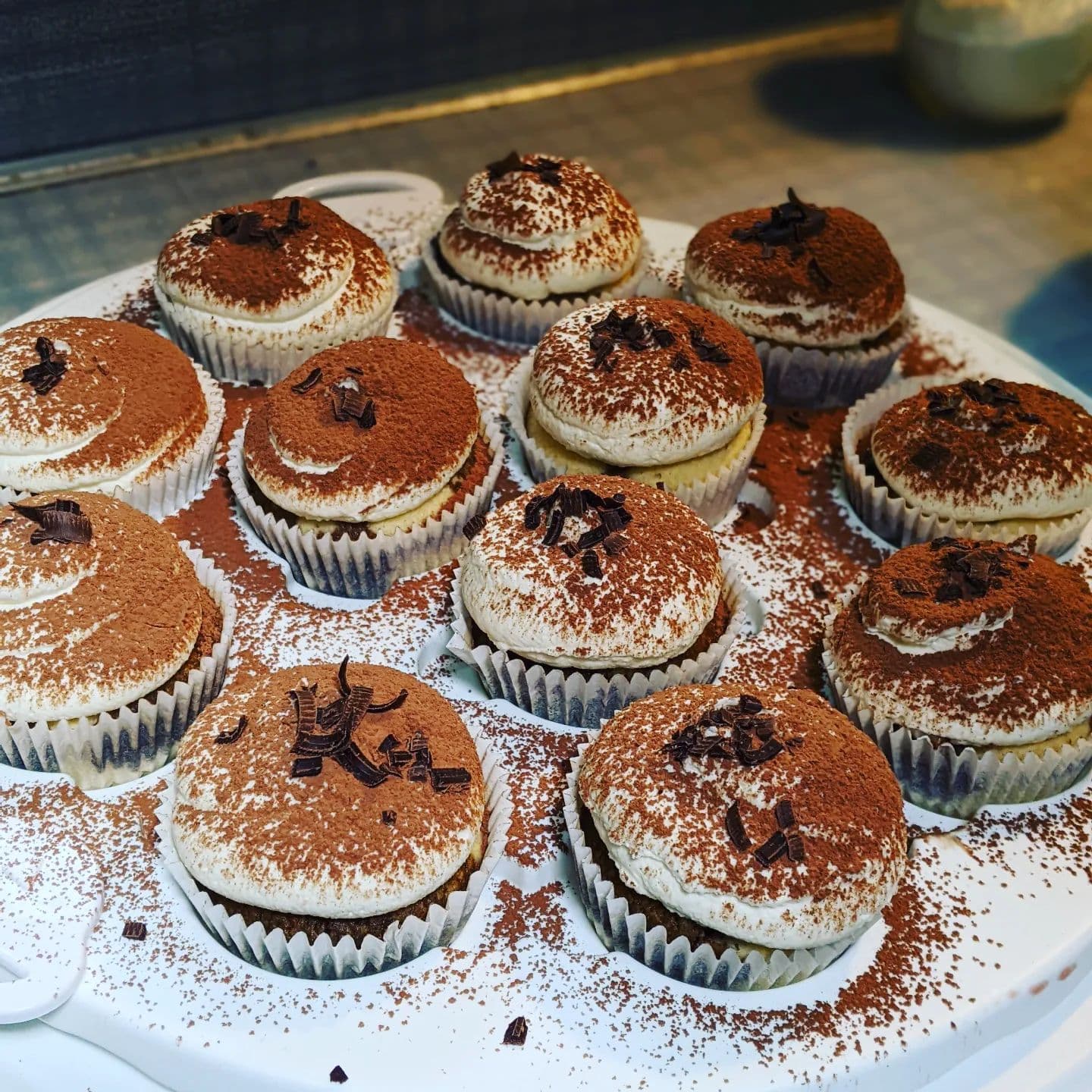 Tiramisu cupcakes with white frosting, cocoa powder, and chocolate shavings on a white tray.